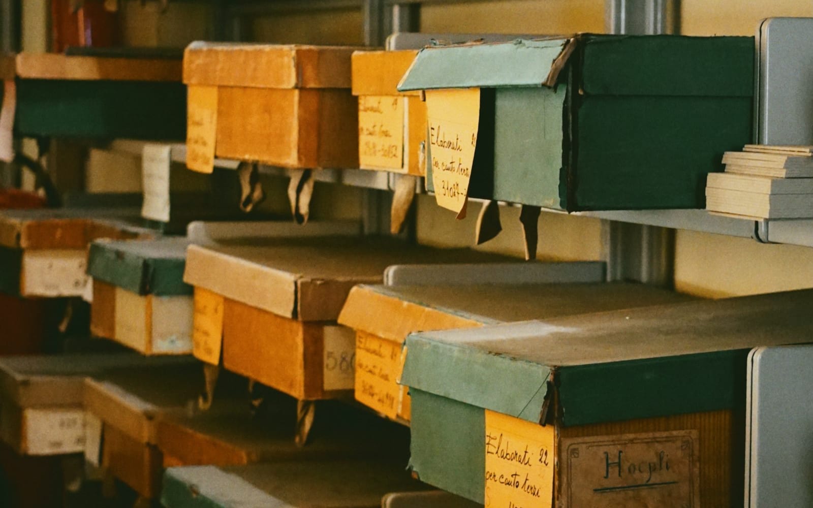 Rows of old cardboard archive boxes on metal shelves, each with a handwritten paper tag hanging from its front
