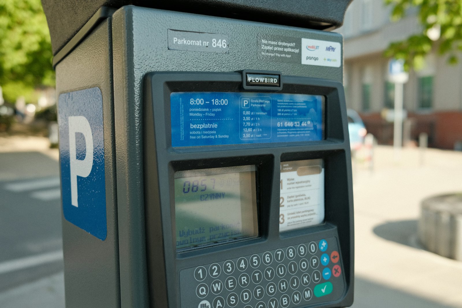 A street parking meter in Poznan with its hourly rates, a minimum charge and a keypad, a green tree and a building blurred behind it